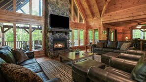A log cabin interior with a stone fireplace, leather furniture, and large windows offering views of the surrounding forest. - Premium Hocking Hills Group Lodge 8BR 5.5Ba Hot Tub, Pond, Starlink, Game Room (Logan)