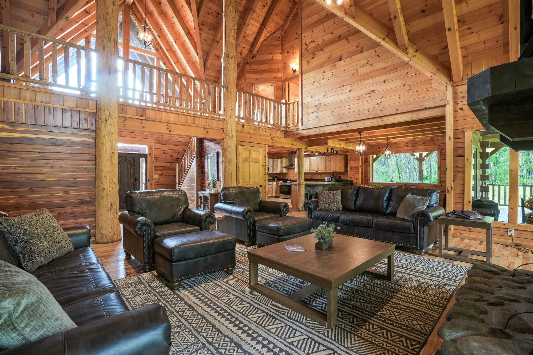 Interior espacioso de una cabaña de madera con techo alto, zona de estar central con sofás de cuero, comedor y cocina.