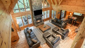 A spacious wooden cabin with a stone fireplace, leather sofas, and large windows offering a view of the outdoors. - Premium Hocking Hills Group Lodge 8BR 5.5Ba Hot Tub, Pond, Starlink, Game Room (Logan)