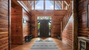 A wooden cabin interior with a central door leading outside, a patterned rug on the floor, and wooden walls. - Premium Hocking Hills Group Lodge 8BR 5.5Ba Hot Tub, Pond, Starlink, Game Room (Logan)