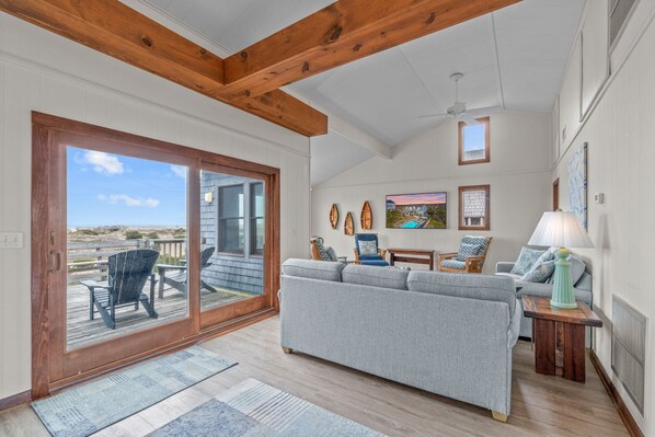 A spacious living room with a large gray sofa, wooden ceiling beams, a view to the outside, and a small table with a lamp.
