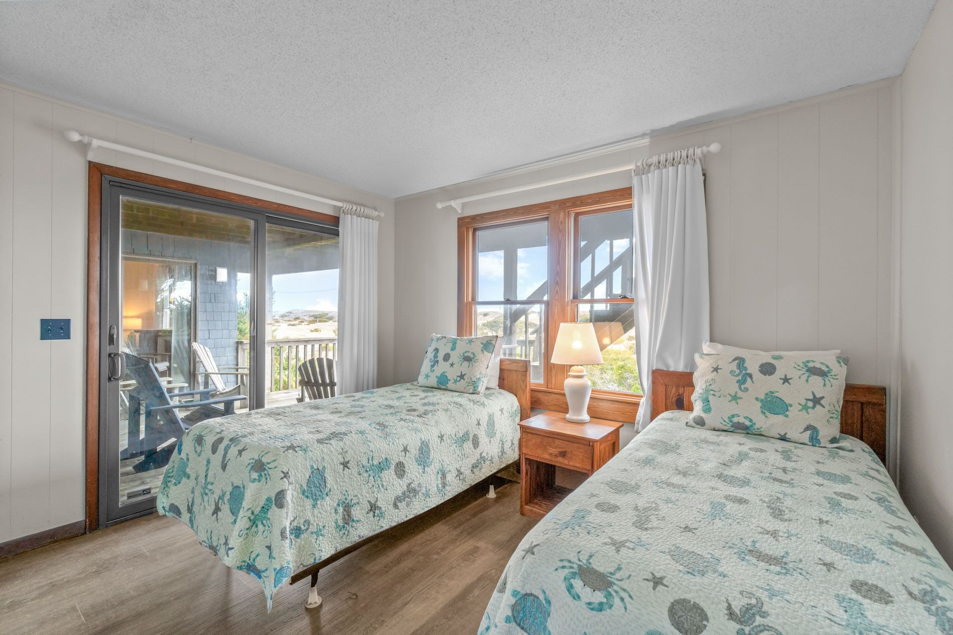 A bedroom with two beds, a wooden nightstand, a lamp, and a view of the outdoors through a sliding glass door.