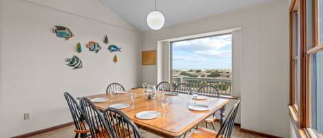 Un coin repas avec une table en bois dressée pour quatre personnes, entourée de chaises, une grande fenêtre donnant sur la plage et des décorations murales représentant des poissons.