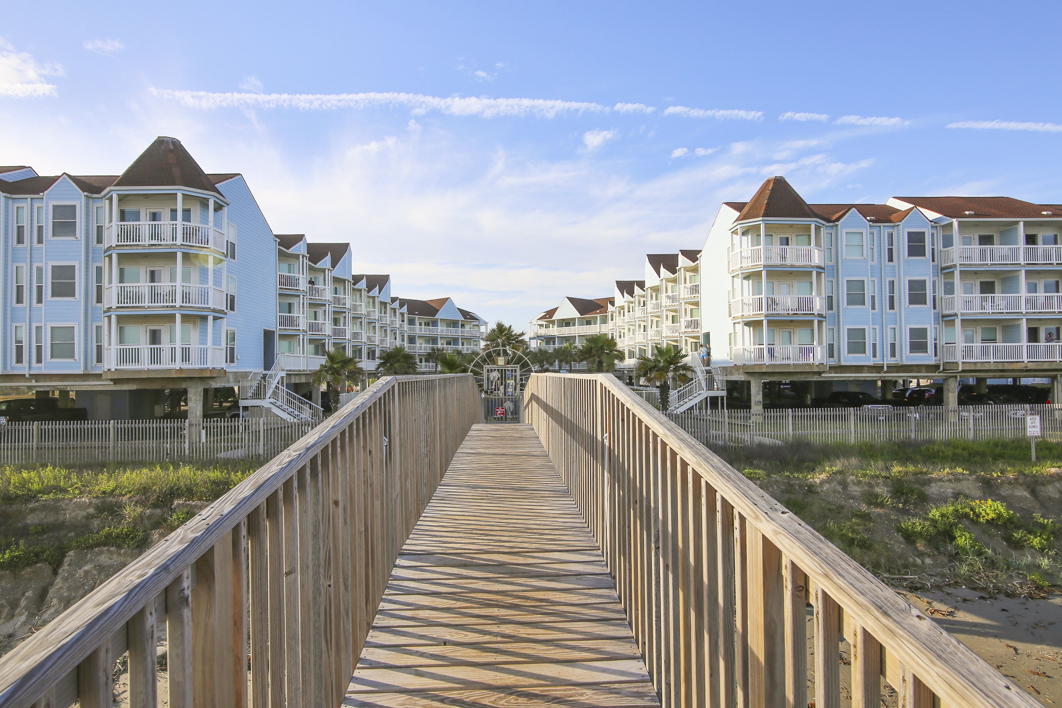 A wooden boardwalk leading to a row of beachfront condominiums with balconies and a clear sky.