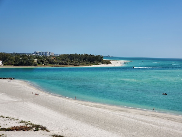 Uma praia com águas turquesa cristalinas, um areal extenso e prédios e árvores ao fundo.
