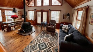 A cozy cabin interior with a wooden floor, a fireplace, a sofa, a dining table, and large windows.