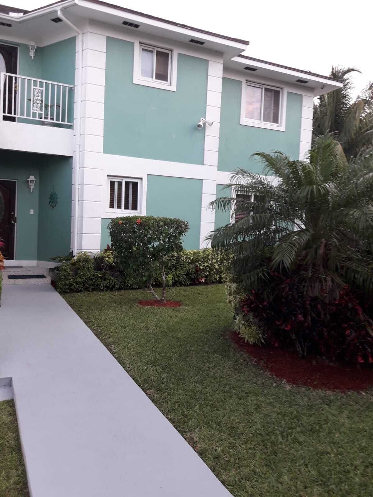 A two-story house with a green and white exterior, a front porch, and a well-maintained lawn with various plants.