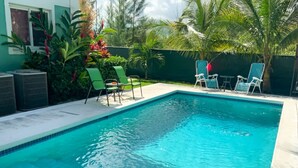 A rectangular swimming pool with clear blue water, surrounded by a concrete deck. There are several lounge chairs and a small table with chairs. The pool is bordered by a low wall with tropical plants and trees in the background. - Cedar Pine Villas - Quiet, 'Luxury & Comfort Rolled Into One' (Nassau)