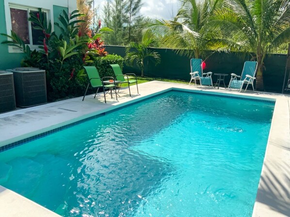 Une piscine rectangulaire à l’eau d’un bleu limpide, entourée d’une terrasse en béton. On y trouve plusieurs chaises longues et une petite table avec des chaises. La piscine est bordée d’un petit muret, et en arrière-plan, on aperçoit des plantes et des arbres tropicaux.