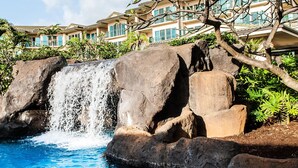 A pool with a waterfall, surrounded by large rocks and a tree. -  Luxury Ocean Front Property!  Ocean& pool view.  B204  (Kapaa)