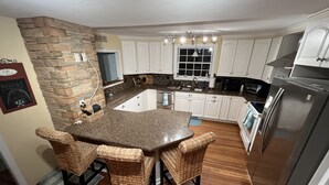 A kitchen with a stone accent wall, white cabinets, a central island with chairs, and a stainless steel refrigerator. - Jamestown: Cozy Coastal Cottage in town near beaches & Newport, dogs OK! (Jamestown)