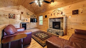 A log cabin interior with a stone fireplace, leather seating, wooden walls, and a ceiling fan.