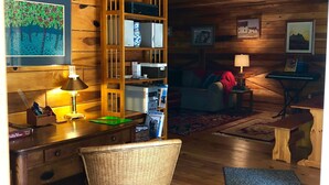 A cozy cabin interior with a wooden ceiling, a desk with a lamp, a wicker chair, and a bookshelf. - Unique farm guest house close to Chattanooga, TN- great for families and dogs (Chickamauga)