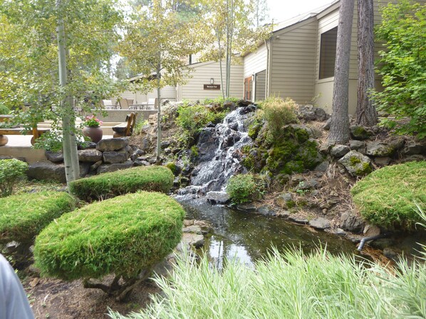 A serene garden with a small waterfall, neatly trimmed bushes, and a stone pathway leading to a building labeled "Broken Top".