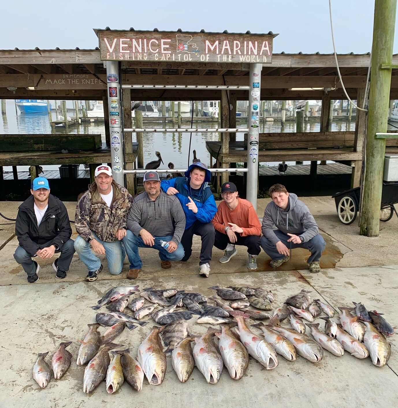 Un groupe de personnes posant devant une structure en bois sur laquelle est affiché un panneau indiquant : « Venice Marina, capitale mondiale de la pêche. »