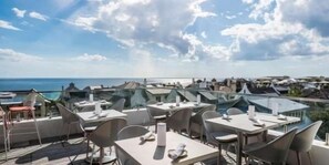 A rooftop terrace with tables and chairs, overlooking a coastal area with buildings and the sea.