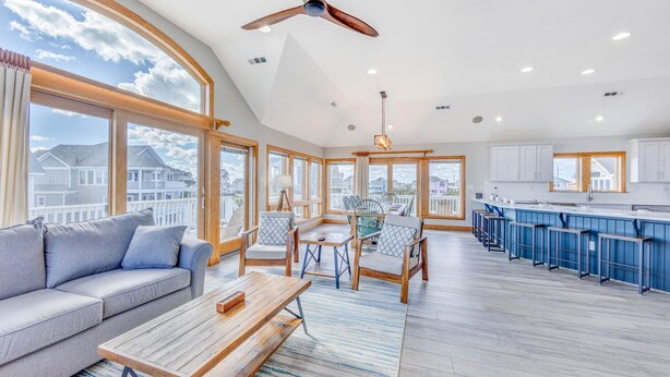 A spacious living room with a large window, a ceiling fan, and a kitchen area with blue cabinets. - Oceanview Dog Friendly Home w/ Pool, Hot Tub, Elevator, Game and Media Rooms (Avon)