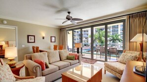 Quarto de hotel com sofá, poltronas, mesa de centro, ventilador de teto e uma janela grande com vista para uma piscina e edifícios.