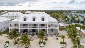 Un edificio grande de varios pisos con fachada blanca, múltiples balcones y ubicación frente a la playa, con tumbonas y palmeras.