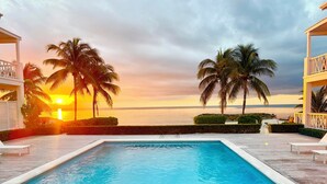 Un área de piscina con vista al atardecer sobre el océano, rodeada de palmeras y una terraza de madera.