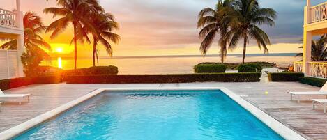A pool area with a view of the sunset over the ocean, surrounded by palm trees and a wooden deck.