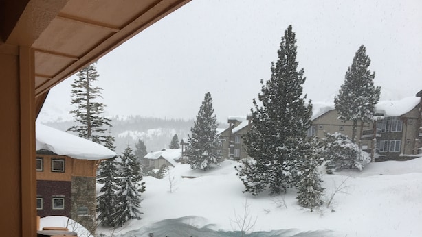 A snowy outdoor scene with a wooden balcony railing, a snow-covered landscape, and a building with a pool in the background.