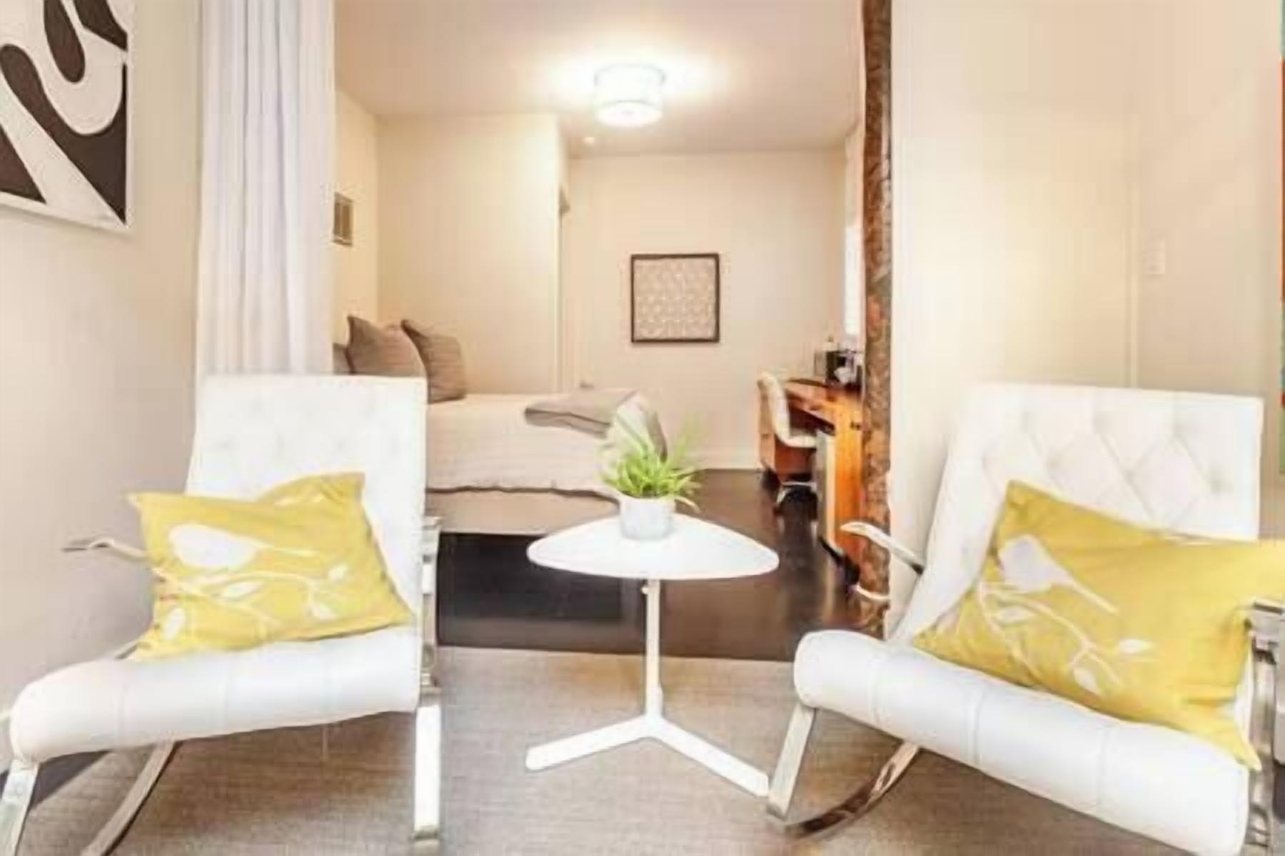 A hotel room with two white armchairs, a small round table with a plant, a bed with a white headboard, and a desk with a chair.