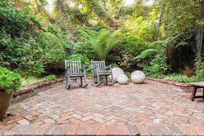 A patio with a brick floor, two wooden rocking chairs, and a variety of plants and trees.