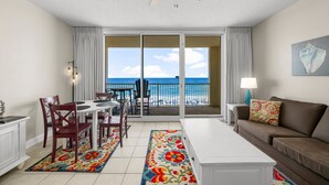A room with a dining table, chairs, a sofa, a ceiling fan, and a view of the ocean through large sliding doors. - 1 Bedroom in Majestic Beach Resort - Low floor, Spacious and bright and beautifully decorated! (Panama City Beach)