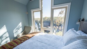 A bedroom with a large bed, a chair, and a view of trees through two windows.