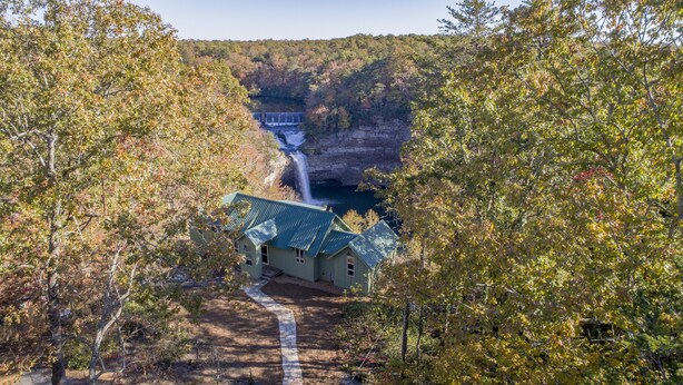 A rustic cabin with a metal roof, surrounded by autumn foliage and a waterfall in the background.
