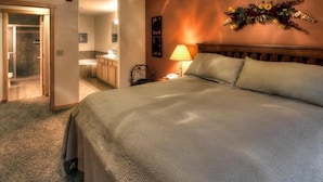 Een hotelkamer met een bed, nachtkastje, lamp, wanddecoratie met bloemen en uitzicht op de badkamer.