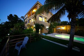 A two-story building with a swimming pool, surrounded by greenery and a wooden fence.