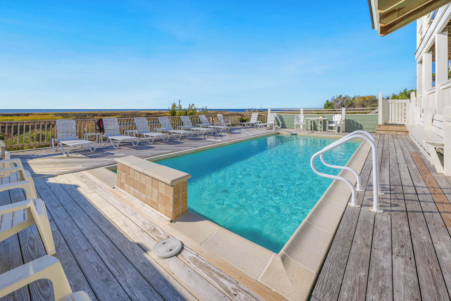A pool area with a wooden deck, white lounge chairs, and a view of the ocean.