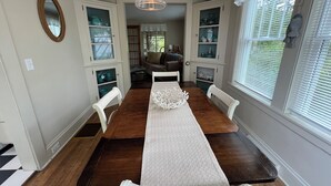 A dining area with a long wooden table, white chairs, a hanging light fixture, and a view of a living room through an open door.
