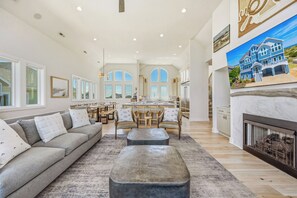 A spacious living area with a sofa, ottoman, dining table, and chairs. A kitchen with a large island and a fireplace is visible. - Premier Oceanfront w/ Pool, Hot Tub, Elevator, Game & Theater Rooms, Tiki Bar (Avon)