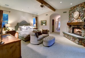 Une chambre spacieuse avec un grand lit, un canapé et un pouf. On y trouve une cheminée en pierre et une vue sur l’extérieur par une fenêtre.