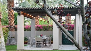 A covered patio with a table and chairs, surrounded by lush greenery and flowering plants.