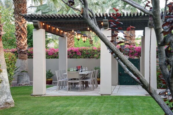 Une terrasse couverte, agrémentée d’une table et de chaises, entourée d’une végétation luxuriante et de plantes en fleurs.