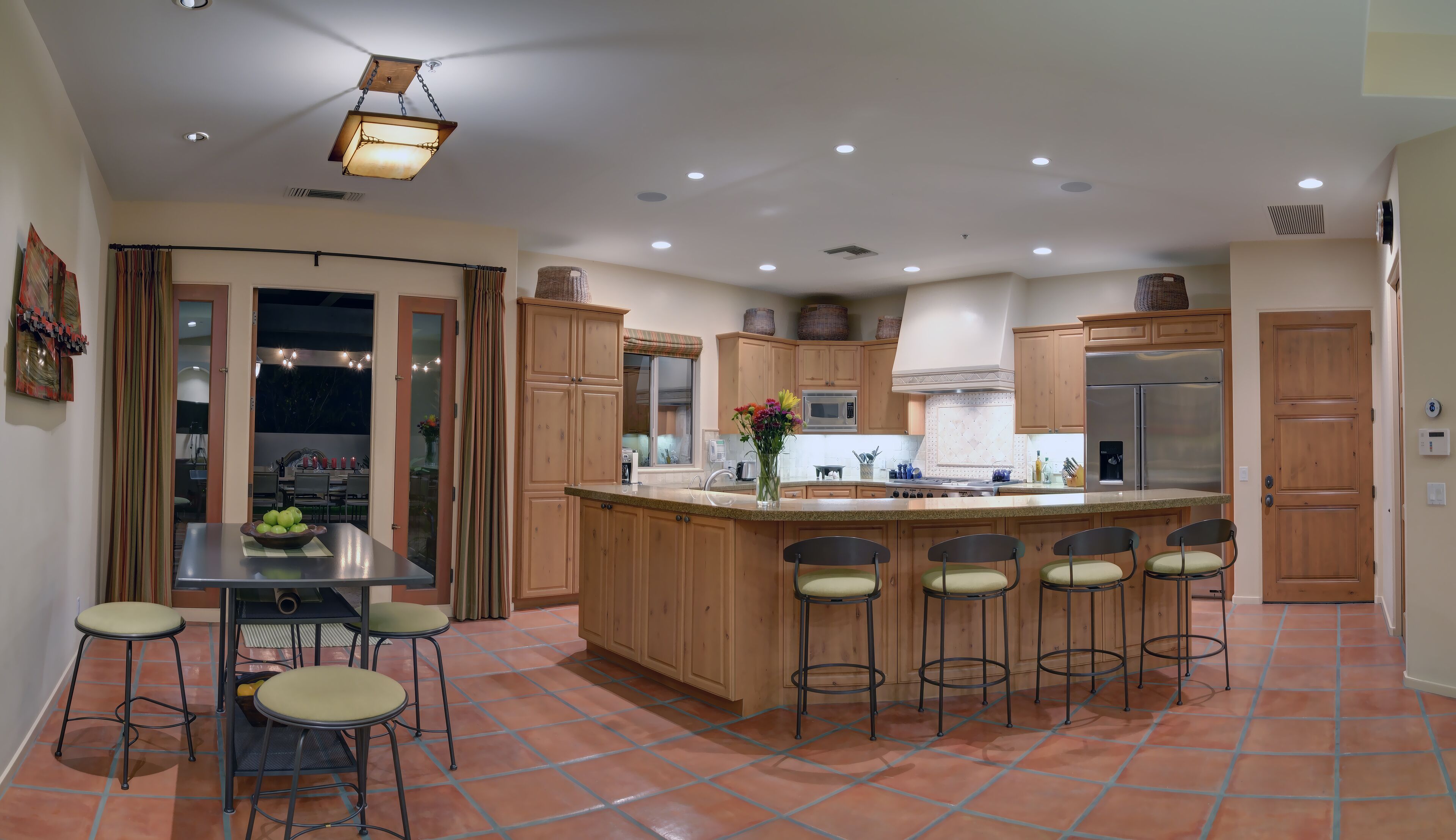 A modern kitchen with wooden cabinets, a central island with bar stools, a stainless steel refrigerator, and a dining area with a table and chairs.