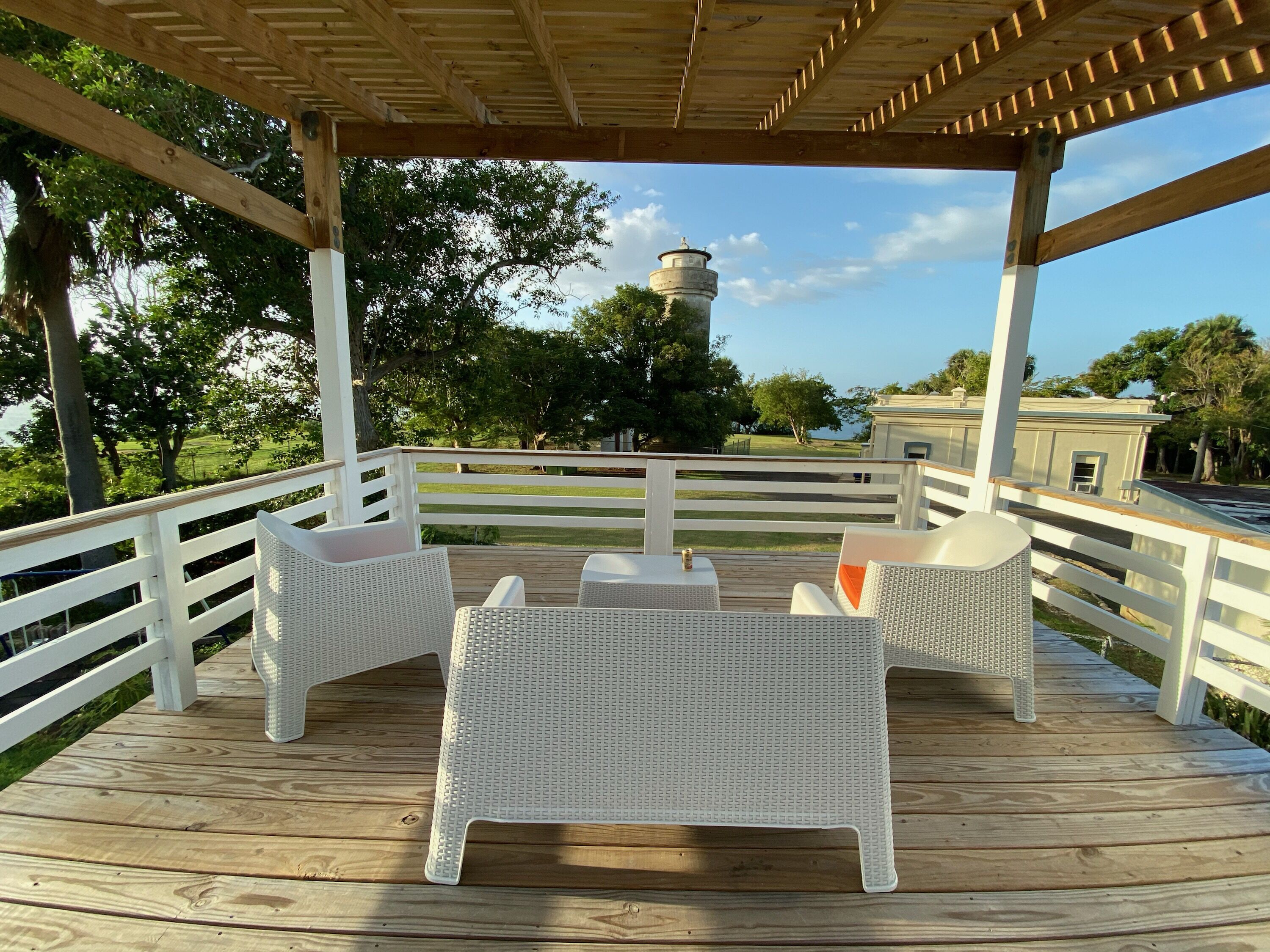 Una terraza techada al aire libre con mobiliario de ratán blanco y vistas a una torre de agua y árboles.
