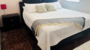 A neatly made bed with a white quilt and two decorative pillows, a dark headboard, a small bedside table with a lamp, and a patterned rug on a dark floor. - El Faro Surf House!!  Less Than 2 mi From Airport And Surf (Aguadilla)