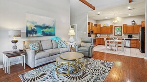 A spacious living room with a gray sofa, a coffee table, and a patterned rug. The kitchen in the background has wooden cabinets and a dining area with white furniture.
