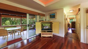 A room with a wooden floor, a TV on a stand, a dining table with chairs, and a view of greenery through large windows.