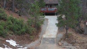 Uma casa com deck vermelho e garagem, cercada por mata densa e um caminho de cascalho.