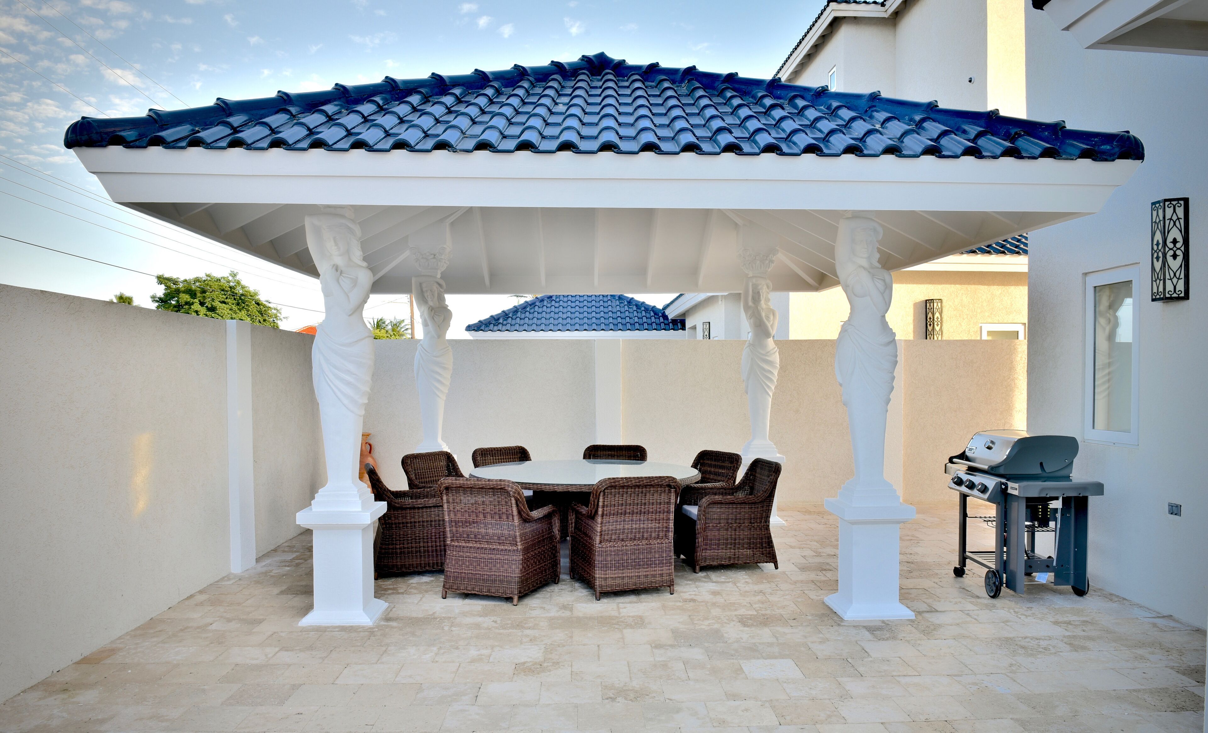 Un patio techado con una mesa redonda y sillas de mimbre, una parrilla y una estructura de columna blanca.