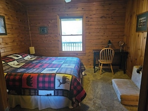 A wooden cabin room with a bed, a desk, a chair, and a window.