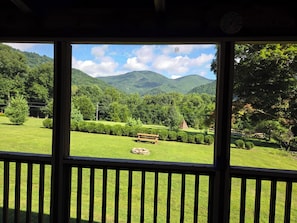 A view through a window with a wooden railing, featuring a grassy area with a bench, trees, and mountains in the background.