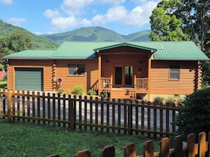 A log cabin with a green roof, a front porch, and a fenced yard.
