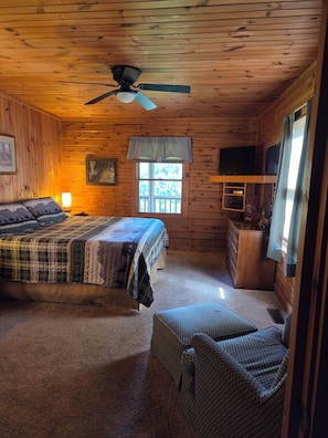 A cabin-style room with a bed, a chair, a television, and a window with a view of trees.